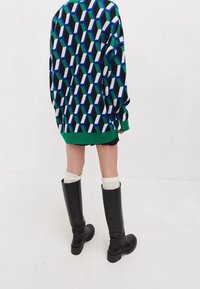 Person som bär en tröja med geometriskt mönster i grönt, blått, svart och vitt, kombinerat med svarta knähöga stövlar och krämfärgade strumpor, stående på vit golv.