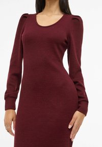 Vestido burdeos acanalado con mangas largas, diseño ajustado y acentos de hombreras abullonadas. Textura suave con escote redondo.