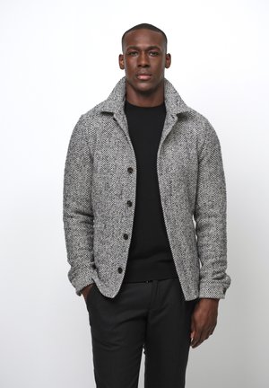 Houndstooth patterned jacket in gray and black, featuring a collar, button front, and textured fabric. Worn over a black sweater.