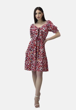Robe fleurie rouge avec des manches courtes bouffantes, détail noué à l'avant et taille cintrée, présentant des motifs floraux multicolores sur un tissu blanc.