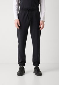 Black tapered joggers with elastic waistband and drawstring, featuring elastic cuffs. Worn with black sneakers, on a neutral background.