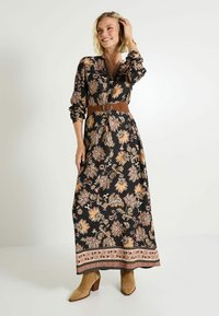 Robe maxi en tissu noir avec un motif floral aux tons dorés et pêche, manches longues, et une ceinture marron à la taille. Bottes à talons marron en dessous.