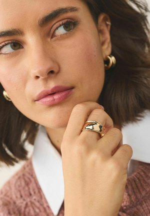 Junge Frau mit kurzen dunklen Haaren, die goldene Creolen und einen breiten, polierten goldenen Ring trägt, ihre Hand in der Nähe ihres Mundes ruhend.