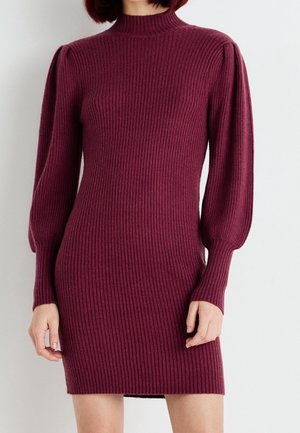 Femme portant une mini-robe en maille côtelée bordeaux à manches longues, épaules bouffantes et col montant.