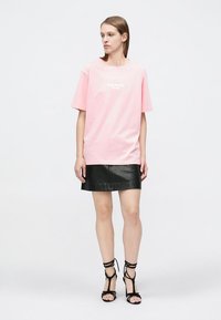 T-shirt en coton rose à manches courtes avec texte blanc, associée à une mini-jupe en cuir noire et des talons noirs à lanières.