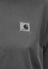 Γκρι ύφασμα με ένα μικρό τετράγωνο λογότυπο Carhartt ραμμένο στην περιοχή του στήθους ενός ενδύματος.