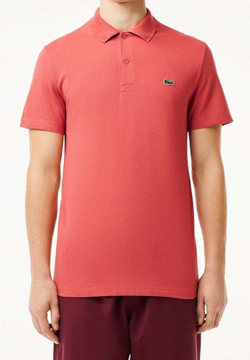 Röd bomullspolo med korta ärmar, treknäppning och en liten grön logotyp på vänster bröst. Slät textur och figursydd design.