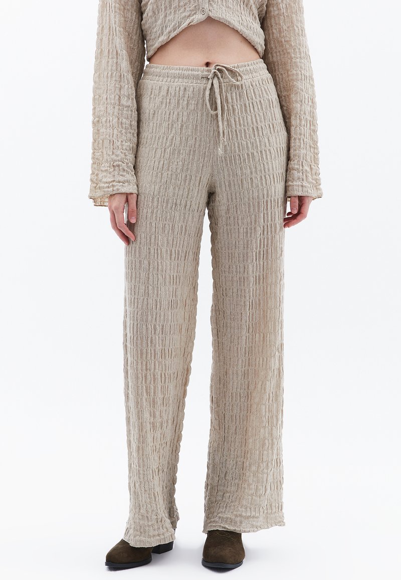 Lichtbeige, gestructureerde, loszittende broek met een trekkoord in de taille en lange, brede benen. Het materiaal lijkt gekreukt met een minimaal patroon.