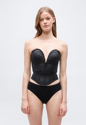 Kvinde med langt blondt hår iført en sort læderkorsette uden stropper og sort højtskåren undertøj mod en ensartet lysegrå baggrund.