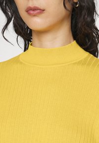 Žlutý žebrovaný rolákový svetr s přiléhavým střihem a vysokým límcem, s měkkou texturou a bezešvou konstrukcí.