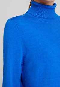 Sininen poolska, pehmeästä neuloksesta, jossa on ribattua kaulusta ja siistejä saumoja, entistä tiiviimpi malli, joka korostaa siluettia.