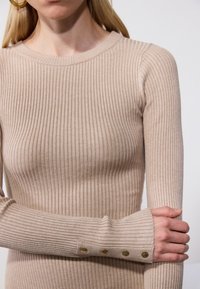 Femme portant un haut beige côtelé à manches longues avec des boutons dorés sur le poignet, tenant un bras croisé sur son corps sur un fond neutre.