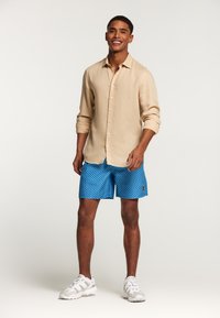 Camicia di lino beige con bottoni, abbinata a pantaloncini blu con un motivo geometrico. Il modello indossa scarpe da ginnastica bianche.