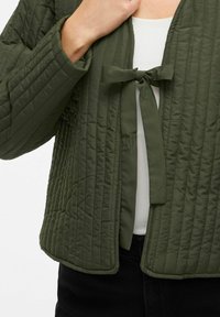 Close-up van een persoon die een gewatteerde olijfgroene jas met een stoffen strik over een witte top en zwarte broek aanpast.