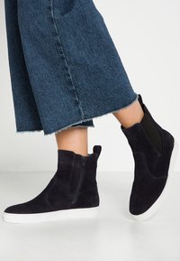 Bottines en suede bleu marine avec fermeture éclair sur le côté et accents élastiques, présentant un dessus texturé et une semelle en caoutchouc blanc.