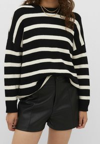 Femme portant un pull à rayures noires et blanches, un short en cuir noir et un collier chaîne doré, se tenant devant un fond uni.
