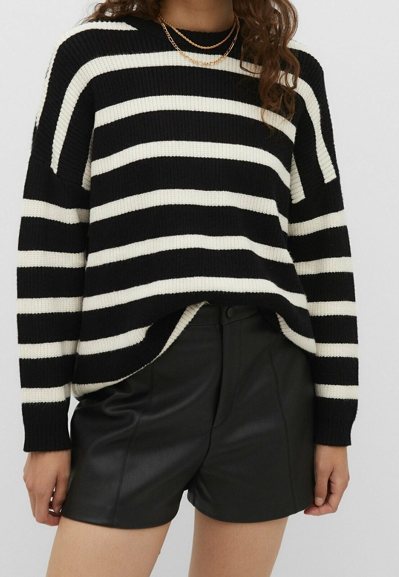 Femme portant un pull à rayures noires et blanches, un short en cuir noir et un collier chaîne doré, se tenant devant un fond uni.