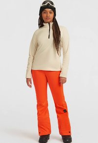 Beige'i värvi võtmega fliis, erksad oranžid lumepüksid, mustad saapad, must beanie ja suusakiivriga naine, seismas valge tausta ees.