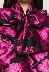 Svart tyg med stort, glänsande magentafärgat blommönster, knutet i en voluminös rosett vid halsringningen på plagget.