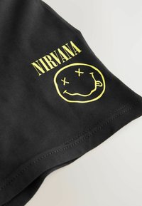 T-shirt en coton noir avec le texte "NIRVANA" en jaune et un graphique de visage souriant avec des X pour les yeux et une langue tirée, à manches courtes.