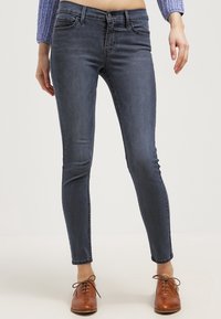 Jeans skinny de color azul oscuro con una cintura de altura media, que incluyen pasadores para cinturón, cinco bolsillos y un desvanecimiento sutil; combinados con zapatos marrones de cordones.