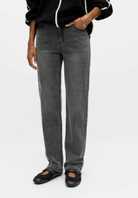 Jeans in denim grigio con un design a gamba dritta, caratterizzati da dettagli di cucitura laterale. Abbinati a calzature nere e a un top nero con accenti bianchi.