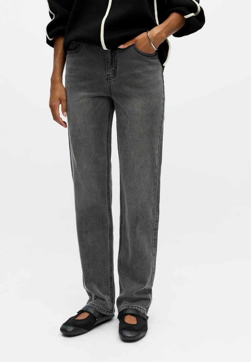 Jeans in denim grigio con un design a gamba dritta, caratterizzati da dettagli di cucitura laterale. Abbinati a calzature nere e a un top nero con accenti bianchi.
