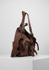 Brun nylonryggsäck med flera fickor, justerbara remmar och nätdetaljer, med en slät yta och rektangulär form.