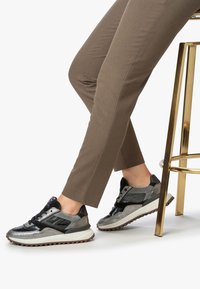 Persona seduta su uno sgabello di metallo dorato, con pantaloni marroni e scarpe da ginnastica casual nere, grigie e argentate con suole testurizzate.
