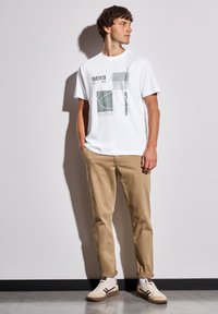 Bílý bavlněný tričko s geometrickým grafickým potiskem v zelené a černé barvě, zkombinované s béžovými kalhotami a krémovými teniskami. Jednoduchý design.