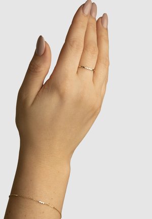 Goldring mit gepunktetem Muster, der an einer Hand mit einem goldenen Armband präsentiert wird. Die Hand zeigt leicht sommersprossige Haut und einen nude-farbenen Nagellack.