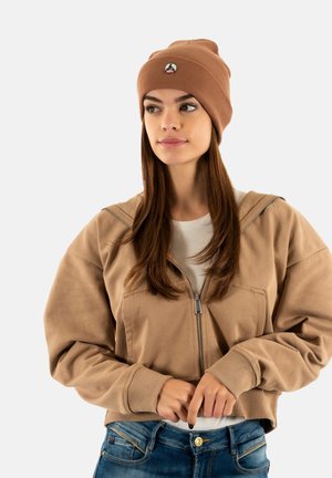 Young woman wearing a tan beanie, beige zip-up sweatshirt, white shirt, and blue jeans, looking slightly to the side with hands near her waist.