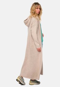 Long cardigan beige avec une capuche, texture tricotée, fentes sur les côtés et coupe décontractée porté sur un haut turquoise avec un jean bleu clair et des baskets grises.