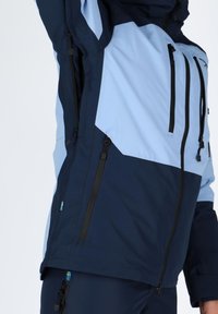 Niebieska i granatowa wodoodporna kurtka z geometrycznym wzorem, wieloma zamykanymi na zamek kieszeniami oraz regulowanymi mankietami. Teksturowana tkanina zapewniająca trwałość.