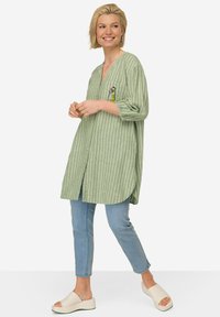 Camicia lunga a righe verdi con scollo a V e dettagli della tasca, abbinata a jeans azzurri chiari e scarpe bianche da indossare facilmente. Tessuto morbido.
