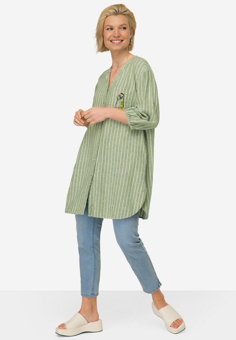 Camicia lunga a righe verdi con scollo a V e dettagli della tasca, abbinata a jeans azzurri chiari e scarpe bianche da indossare facilmente. Tessuto morbido.