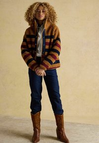 Brūna un tumši zilā svītraina flīsa jaka ar pogām, apvienota ar tumšiem džinsiem un brūniem kovboju zābakiem ar dekoratīvu izšuvumu.