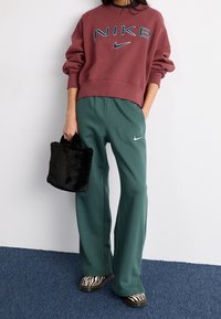 Osoba v maroon mikině Nike, zelených teplákách Nike, se zebra vzorem na botách a držící černou chlupatou kabelku.