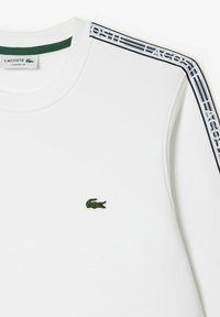 Bílá mikina s zeleným límcem, která má malé zelené logo krokodýla a černobílé pruhované branding Lacoste na ramenou.
