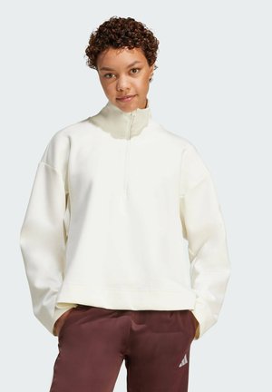 Bijela zip-up dukserica s visokim ovratnikom, oversized kroja i rebrastim manžetama. Uparena s tamnosmešnim hlačama koje imaju detalj s logom.