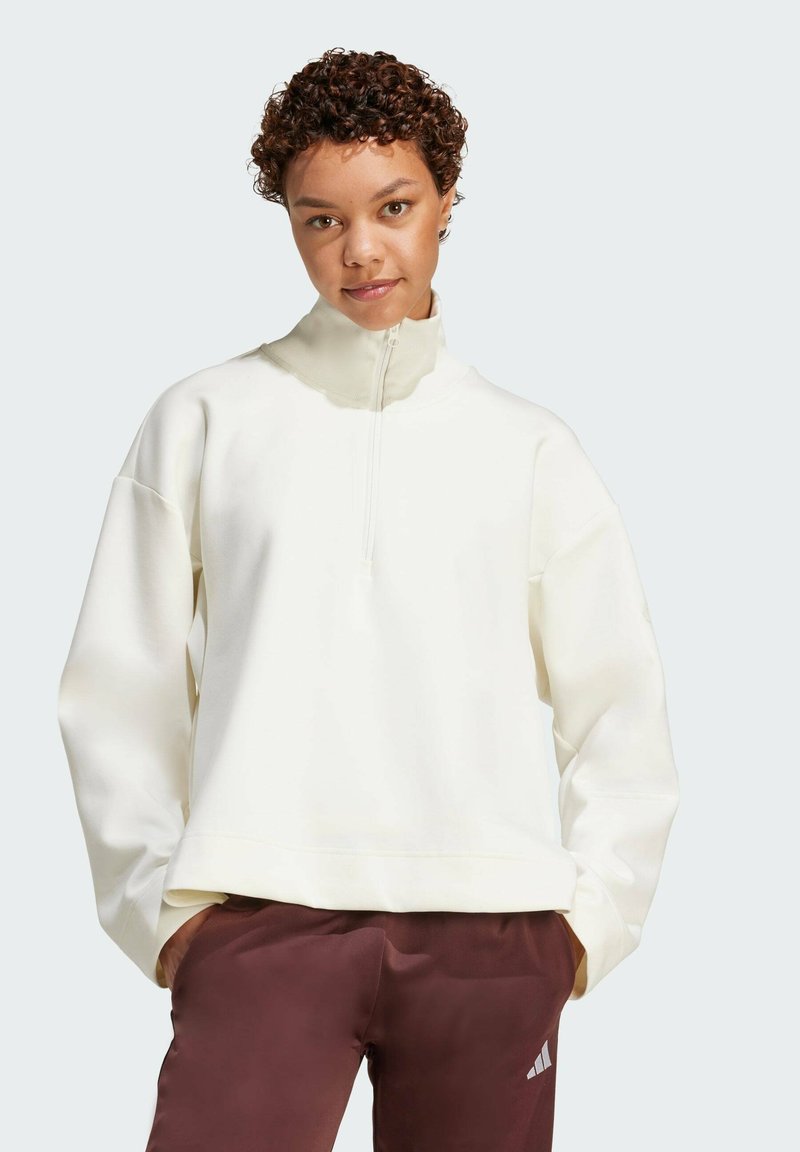 Bijela zip-up dukserica s visokim ovratnikom, oversized kroja i rebrastim manžetama. Uparena s tamnosmešnim hlačama koje imaju detalj s logom.