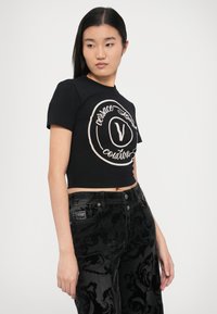 Črna krajšana majica s potiskom kremnega logotipa. Kombinirana s črnimi teksturiranimi kavbojkami z cvetličnim vzorcem. Ozek kroj, kratki rokavi.