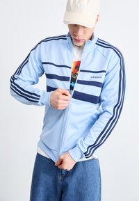 Világoskék, cipzáros dzseki sötétkék csíkokkal és adidas logóval. Magas gallérral és bordázott mandzsettákkal rendelkezik. Egy fehér grafikás póló fölött viselik.