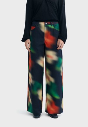 Pantalon à jambes larges avec un motif abstrait multicolore en tons rouge, vert et beige, porté avec un haut noir à manches longues et des chaussures noires.