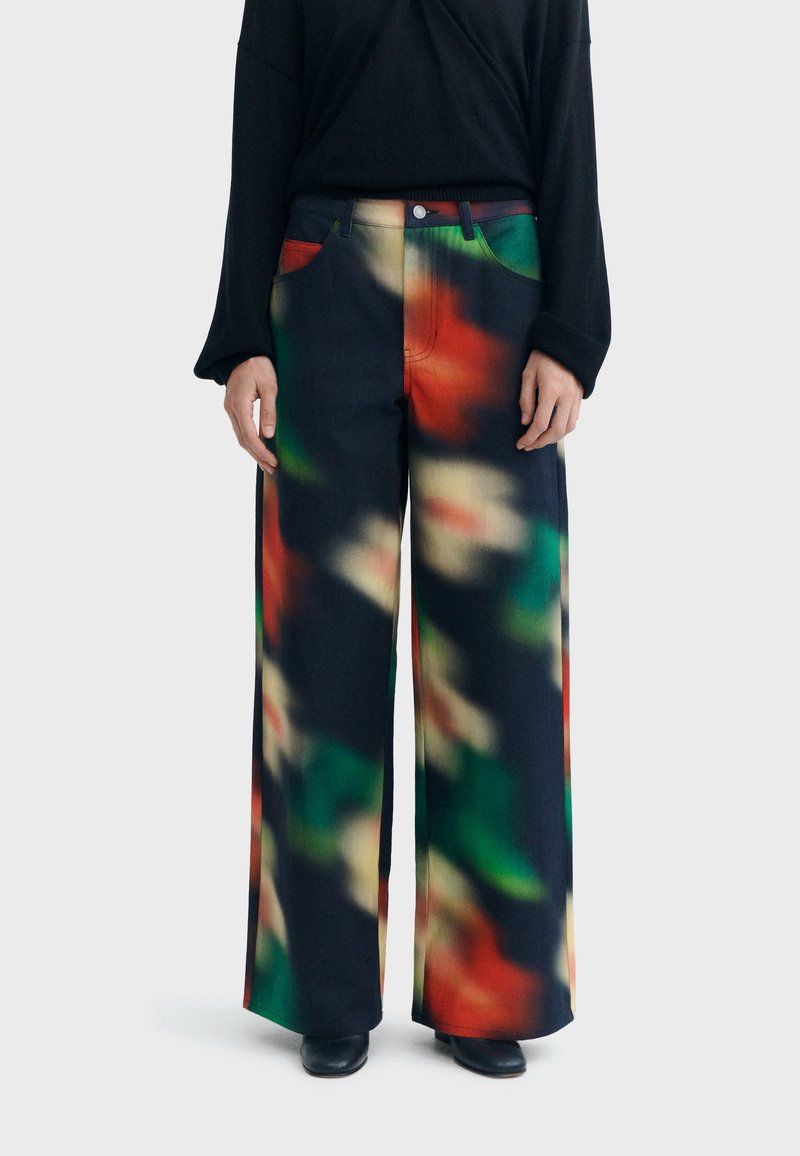 Pantalon à jambes larges avec un motif abstrait multicolore en tons rouge, vert et beige, porté avec un haut noir à manches longues et des chaussures noires.