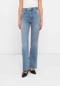 Jeans de denim azul claro com um cós alto, corte de perna reta e design clássico de cinco bolsos. Usados com sapatos de salto preto com ponta fina.