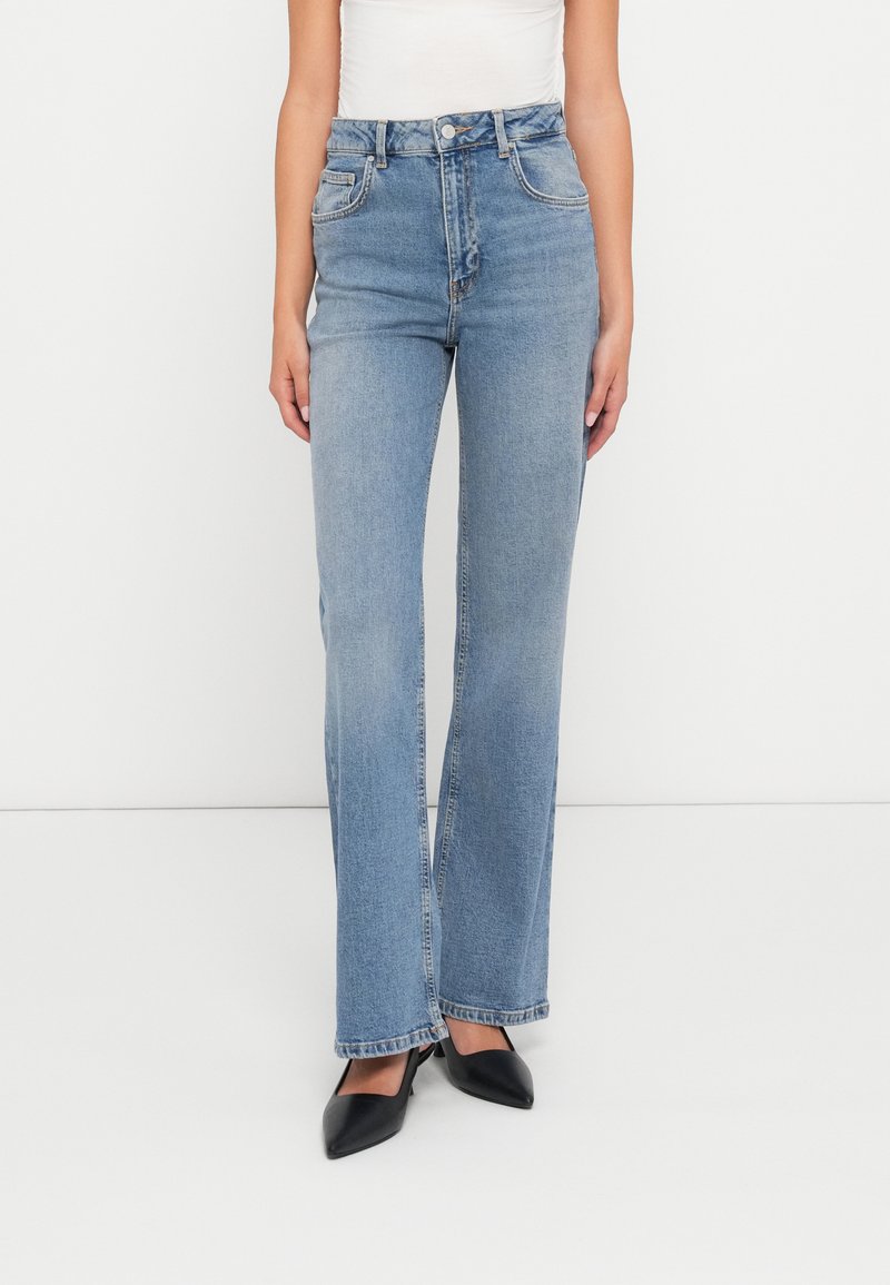 Jeans de denim azul claro com um cós alto, corte de perna reta e design clássico de cinco bolsos. Usados com sapatos de salto preto com ponta fina.