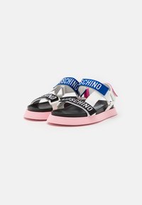 Valkoiset sandaalit, joissa on vaaleanpunaiset pohjat, mustat pohjalliset ja säädettävät siniset ja mustat hihnat, joissa on "Moschino"-brändäys.