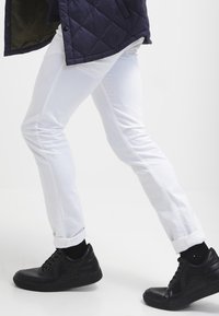Bílé úzké džíny s hladkým povrchem, v slim fit střihu a s ohrnutými lemy, v kombinaci s černými texturovanými nízkými teniskami.