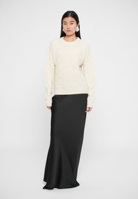Krém színű, texturált kötött pulóver kerek nyakkivágással, amelyet egy elegáns, fekete, padlóhosszig érő selyem szoknyával párosítottak. A modell egy fehér háttér előtt áll.
